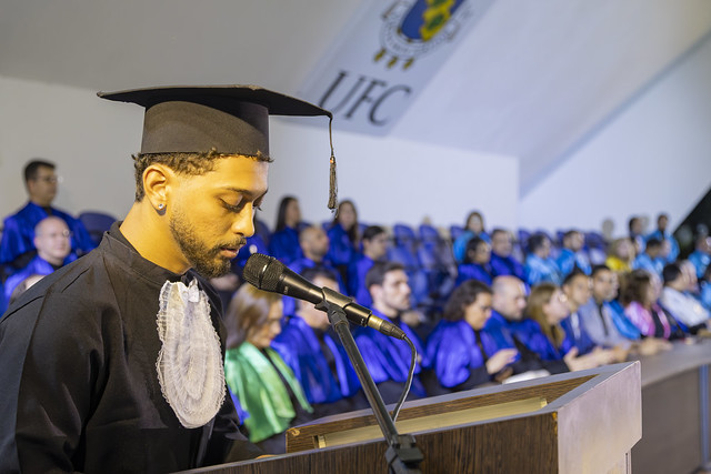 Imagem: um jovem de beca fala em um púlpito. Ao fundo, professores, sentados com veste talar, atentos ao jovem